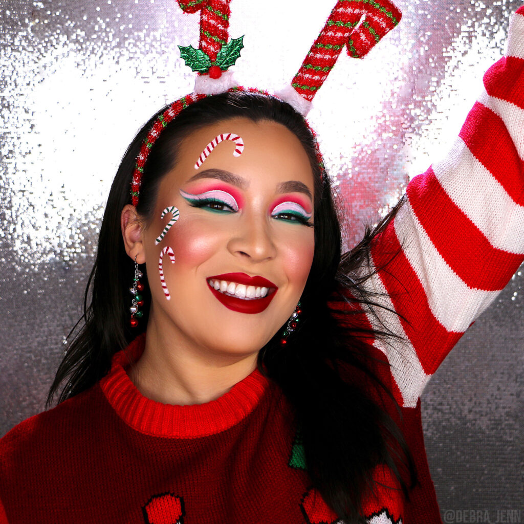 Debra Jenn in red and green eyeshadow with candy canes painted on face, ugly christmas sweater, and candy cane antlers
