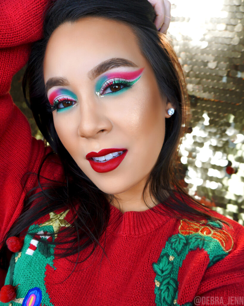 Debra Jenn in green and red eyeshadow for christmas paired with a matte red lipstick and an ugly christmas sweater