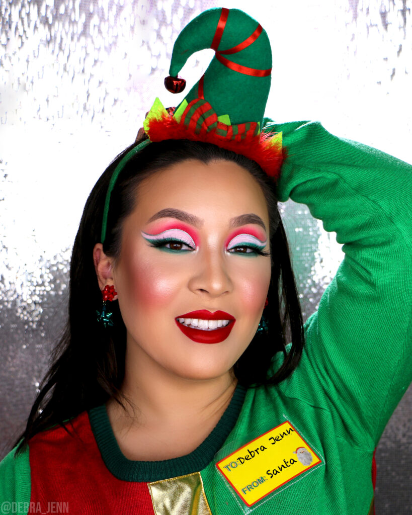 Debra Jenn wearing red and green eyeshadow look with silver eyeliner and red lipstick, wearing an elf hat and gift bow earrings