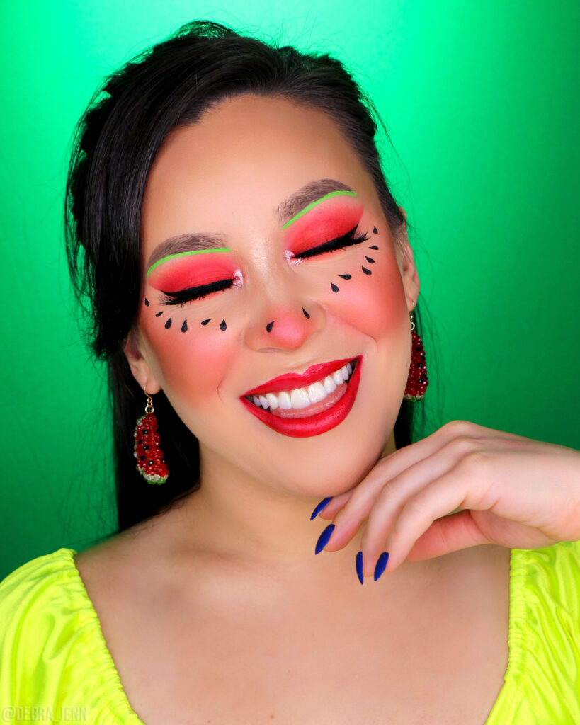 debra jenn in a cute watermelon makeup look with red eyeshadow, red lipstick, green lines under her eyebrows, and watermelon seeds on her cheeks